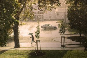 voldsomt regvær. en person med paraply løper langs fortau, en bil står parkert i bakgrunne på asfaltplass. litt plen og trær 